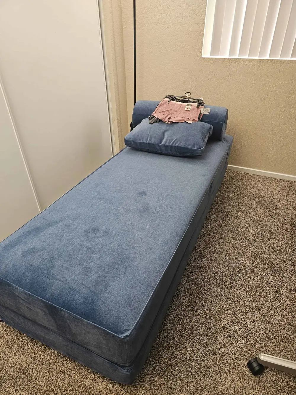 A blue upholstered daybed, reminiscent of swinger sex furniture, with a pillow and a hanger holding a folded pink shirt, sits on a carpeted floor near a beige wall and a window with vertical blinds.