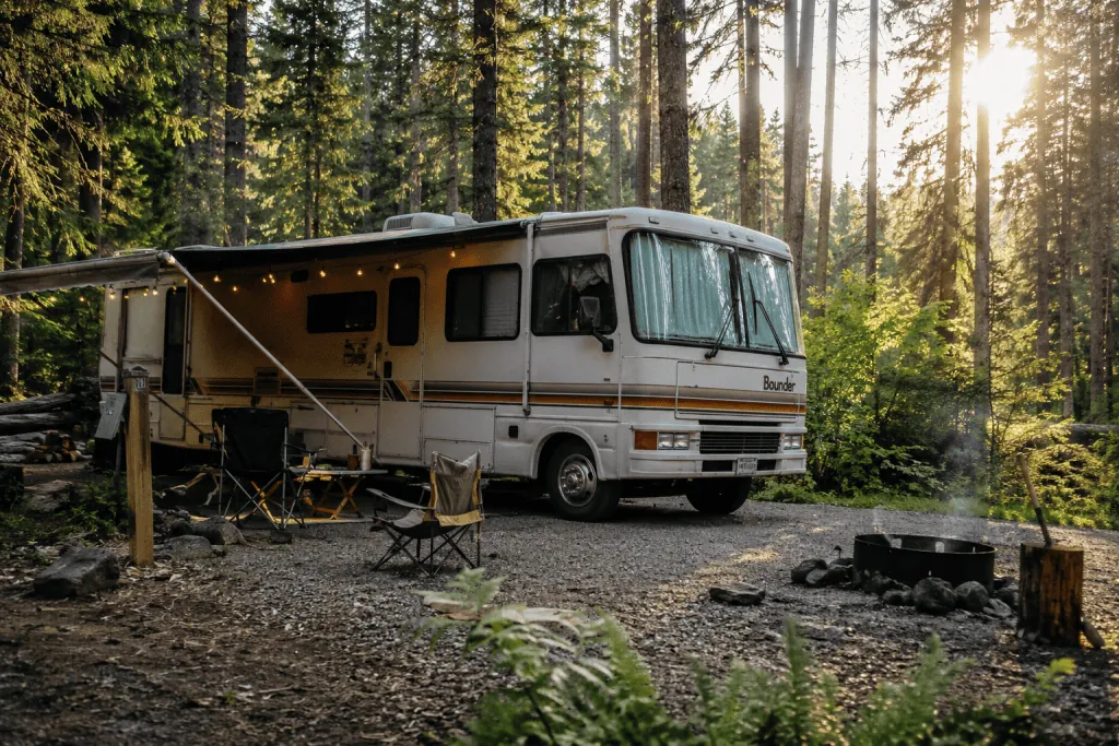 A motorhome parked at a forest campsite with chairs, a fire pit, and trees surrounding the area in sunlight-an inviting spot for couples guide to Naked RVing or an adventurous nudist road trip.