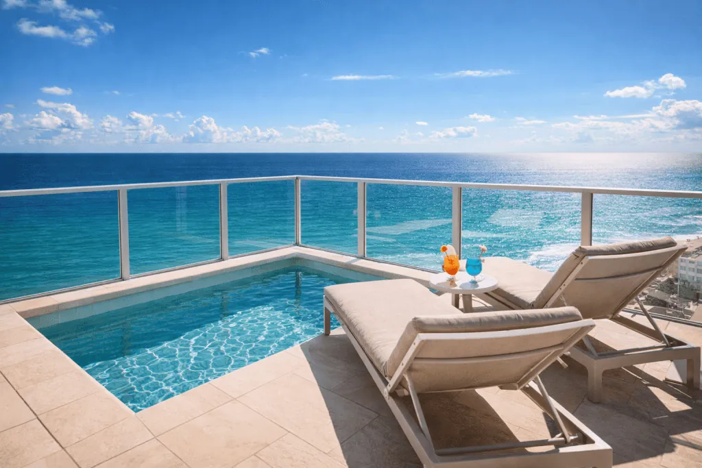 Two lounge chairs and a small table with drinks sit beside a private pool on a balcony overlooking the ocean under a clear blue sky at one of the premier swinger hotels near Miami.