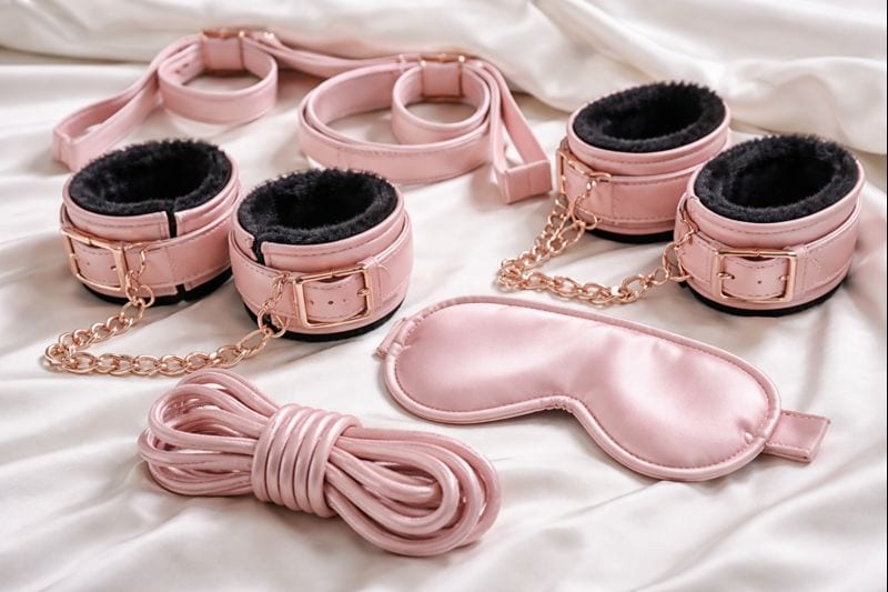 Beginner Bondage Kits That Are Safe And Fun 2 A set of pink faux leather restraints, including wrist and ankle cuffs with gold chains, a blindfold, and a length of soft rope—perfect beginner BDSM kits that are safe and fun, arranged on a white satin sheet.