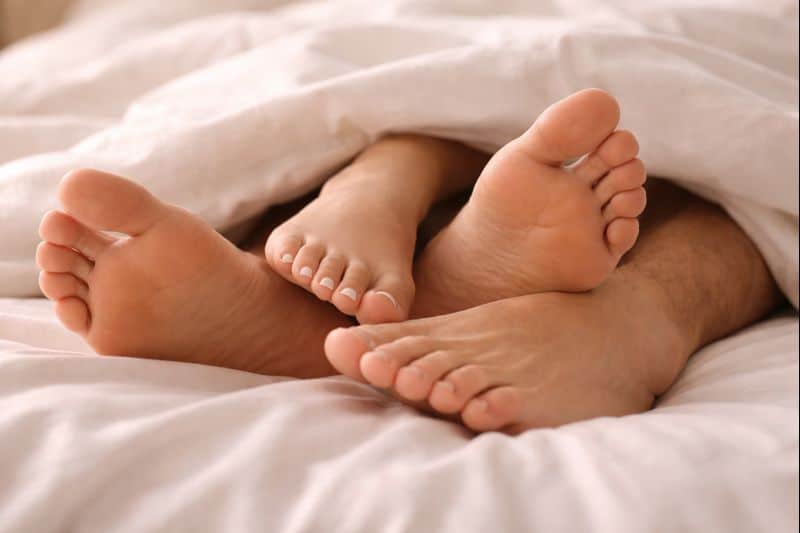 Two pairs of feet, one larger and one smaller, are visible sticking out from under a white blanket on a bed, inviting viewers to explore a foot fetish in a subtle and playful way.
