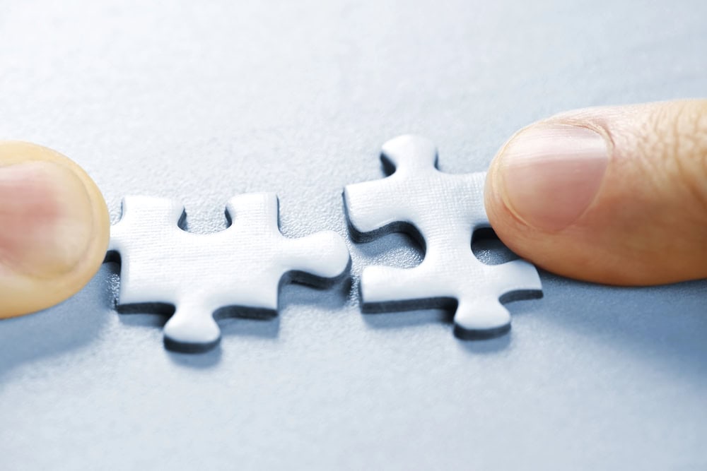 Two fingers holding white jigsaw puzzle pieces, positioned to connect them on a light surface—much like beginner swinger flirting, where lifestyle etiquette guides gentle connections and flirting without pressure.