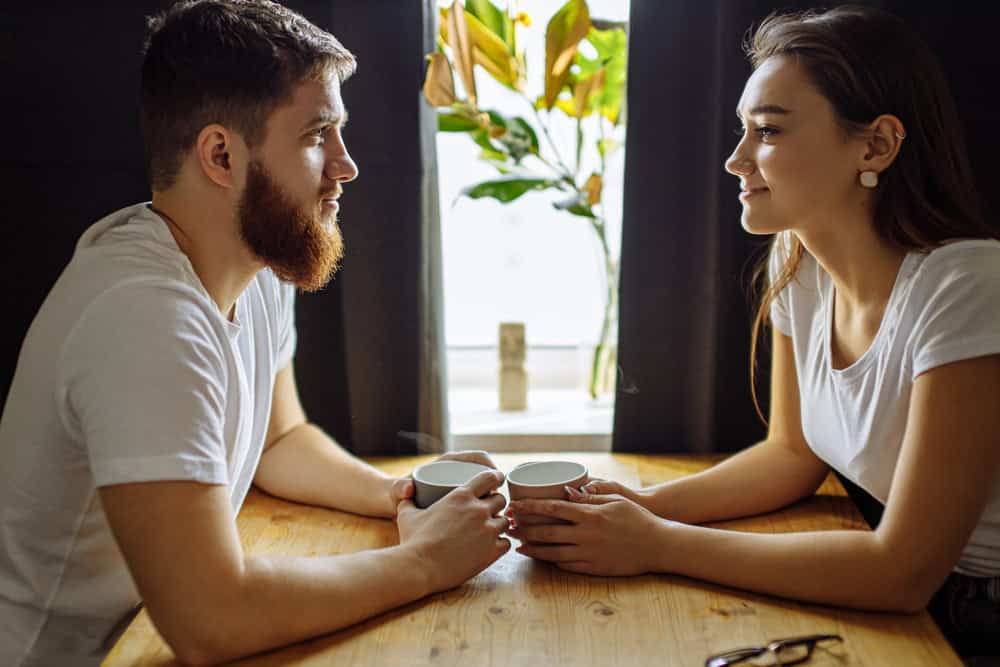 How to Tell Your Partner You Want to Try Swinging 2 A man and a woman sit across from each other at a wooden table, holding mugs and talking, practicing lifestyle etiquette, with a plant and window in the background.