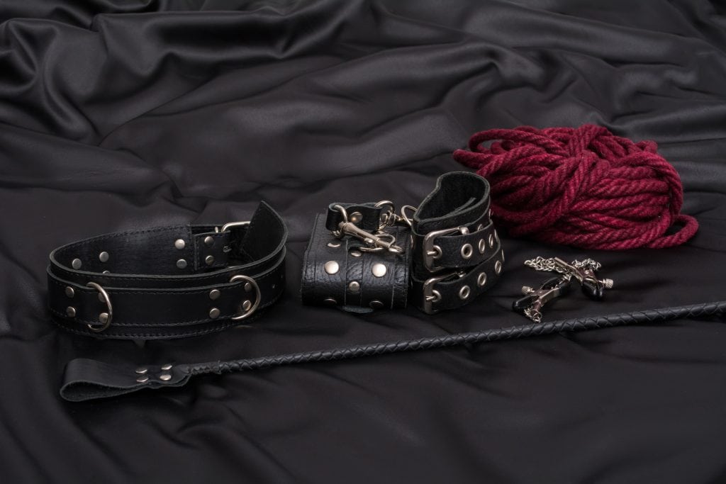 Black leather collar, wrist cuffs, red rope, a small black whip, and a metal cross rest on black satin—a stylish nod to beginner BDSM and consensual kink exploration.
