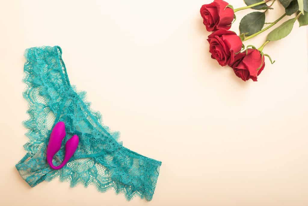 Best Wearable Vibrators for Hotwife Play A pair of teal lace panties and a purple Lovense Lush 3 vibrator, perfect for wearable vibrators for hotwife play, are placed on a light surface next to three red roses.