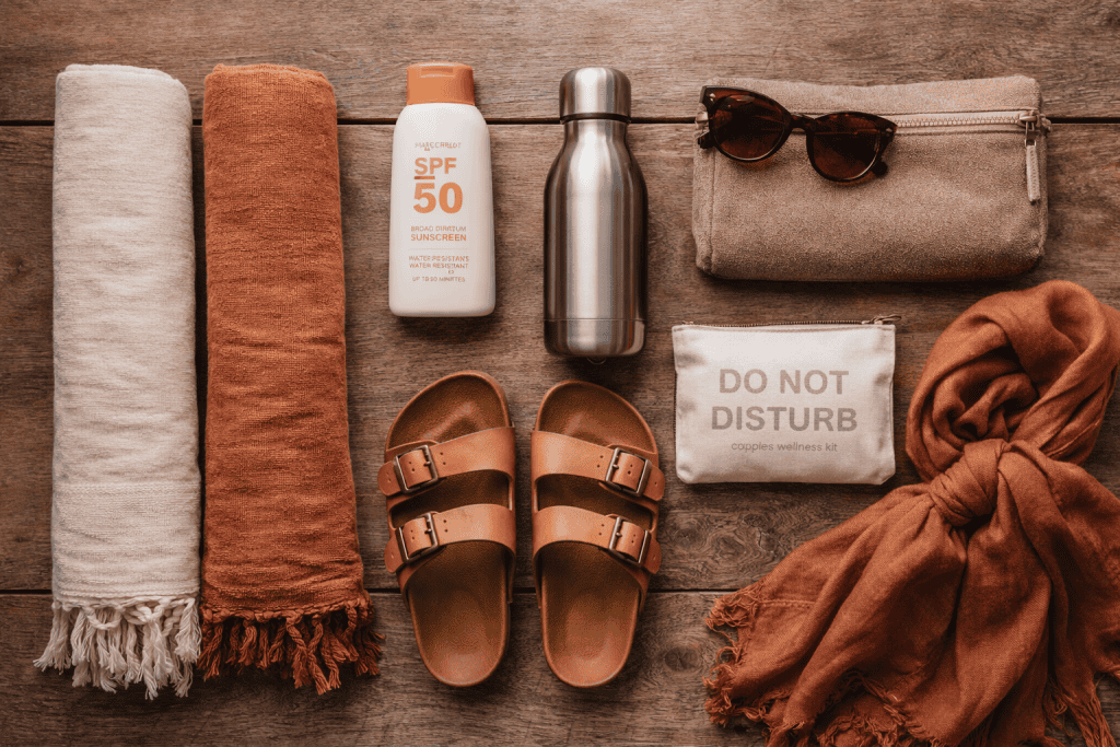 Nudist Camping Packing List. Flat lay of beach essentials, perfect for your nudist camping packing list—two towels, SPF 50 sunscreen, metal water bottle, sandals, sunglasses, pouch, and a scarf on a wooden surface.
