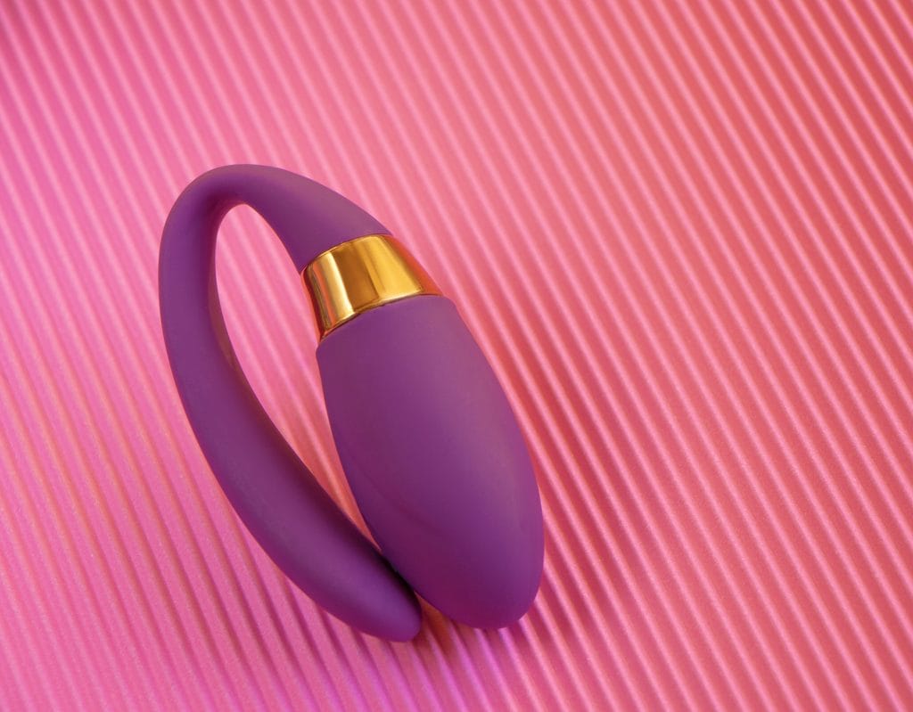 Best Remote Control Toys for Couples purple silicone adult toy with a gold accent, inspired by the best app controlled sex toys for couples, rests on a textured pink striped surface.